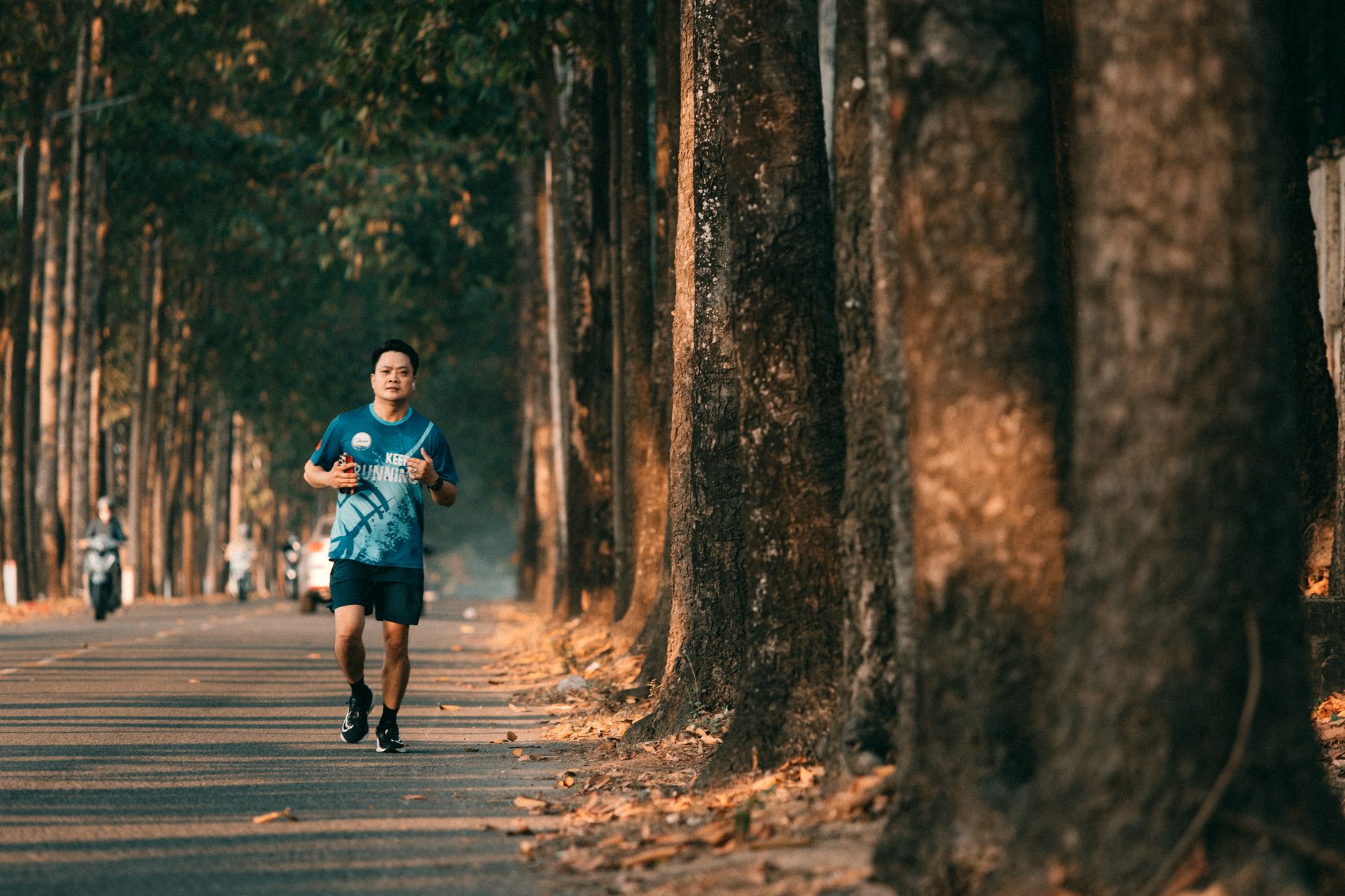 A runner in a blue shirt jogging along a tree-lined road in warm morning light