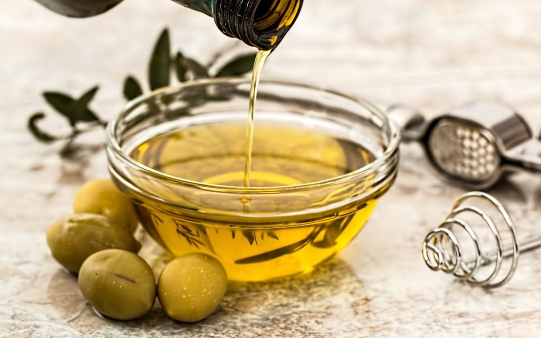 Olive oil being poured into a glass bowl surrounded by fresh olives