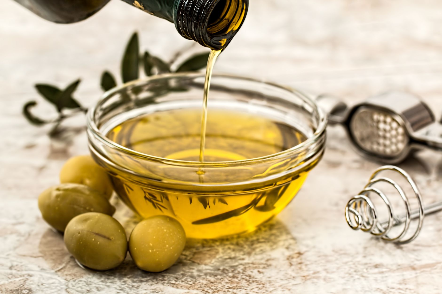 Olive oil being poured into a glass bowl surrounded by fresh olives